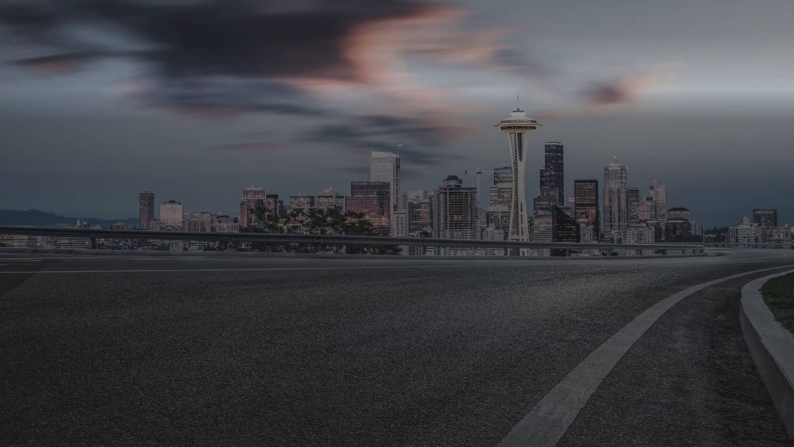 A wide asphalt roadway with a painted edge line, the Seattle skyline beyond it at dusk