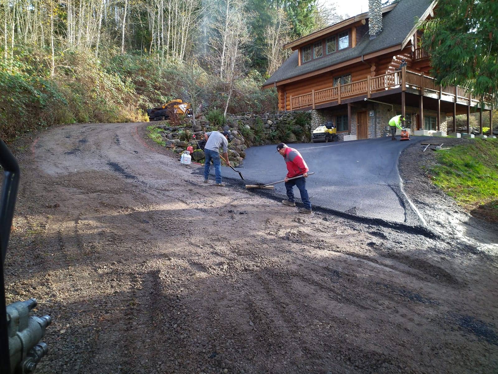 Crew raking fresh asphalt on a wooded hillside driveway